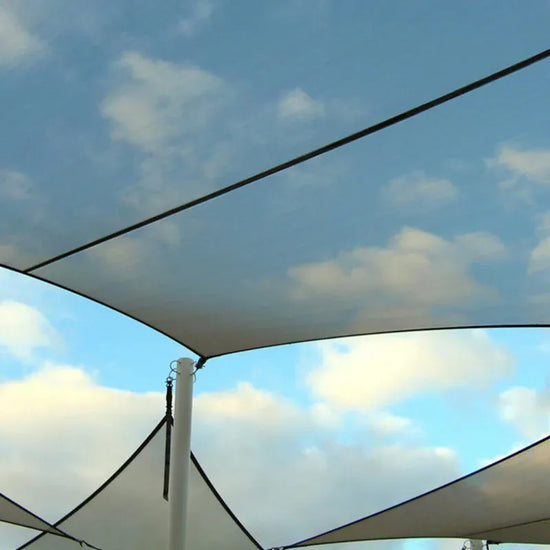 A close-up view of a white tension fabric canopy or shade sail suspended from a white support pole, with blue sky and clouds visible through and beneath the fabric.